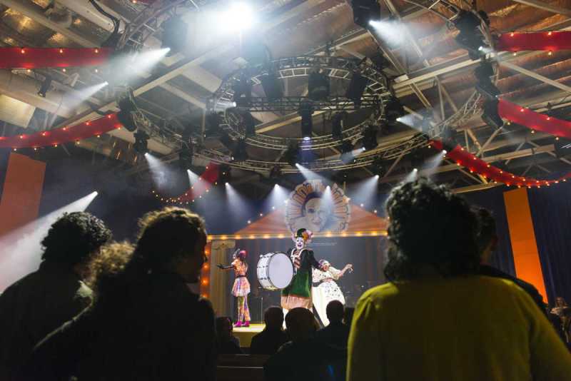 Villagers fill the theater as the troupe begins its commedia dell’arte performance with (l.-r.) Omar Najmi playing Harlequin, Michael Mayes playing Taddeo/Ringmaster, and Lauren Michelle playing Columbina in Boston Lyric Opera’s production of Ruggero Leoncavallo’s Pagliacci.