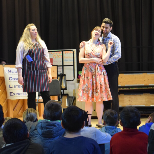 Three BLO Artists are performing The Barber of Seville in a school. A tall blond woman on the left wears a striped apron. To the right, a man gently embraces a woman in a pink dress and their mouths are open in song. Darkened heads and sholders of kids sitting on the floor can be seen in the foreground.