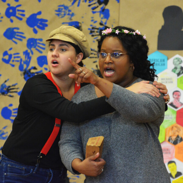 A woman with light brown skin playing Hansel in a hat and red suspenders has her arms around another woman with brown skin and glasses playing Gretel with a flower crown and glasses and a grey top who is pointing. Both singers have mouths open in song. The background is of a childrens art project indicating the picture is taken in an elementary school